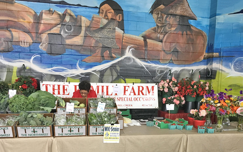 Port Angeles Farmers Market stand with vegetables and flowers in front of a Native American mural