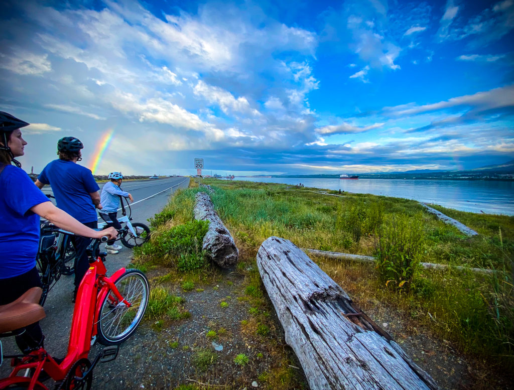 Bicyclists look out over Port Angeles Harbor from Ediz Hook. A rainbow and clouds are in the sky.