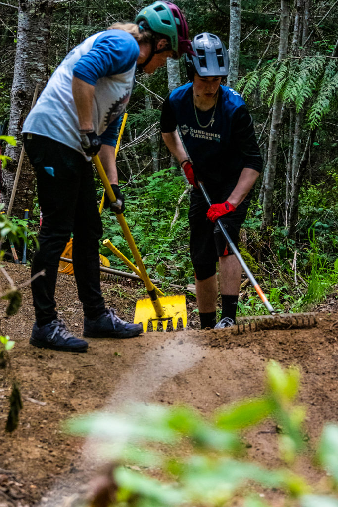 Two people with rakes prepping the dirt on a mountain bike trail in the woods.
