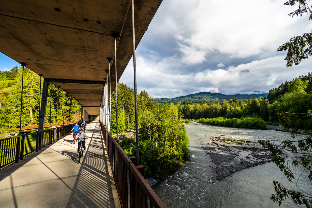 Bicyclists ride on a suspended bridge underneath a road with a river underneath.