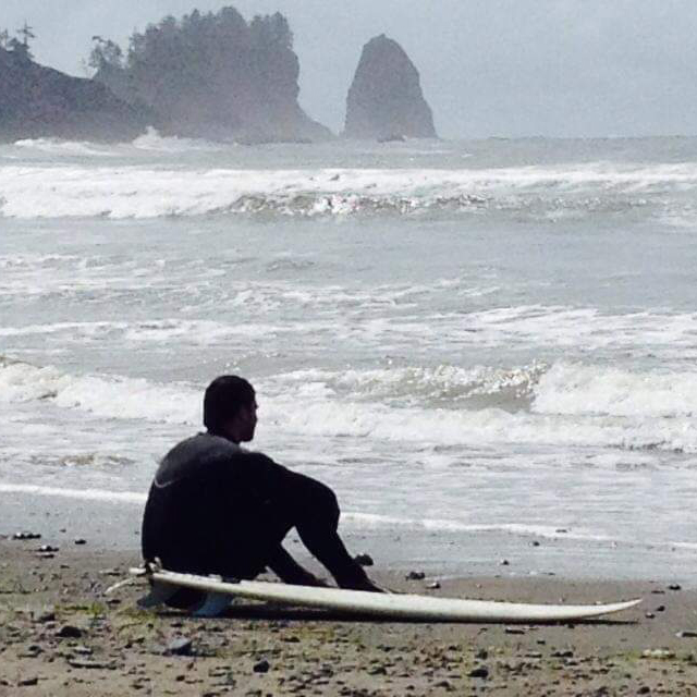 A surfer sits on the beach in a wet suit with seastacks in the background