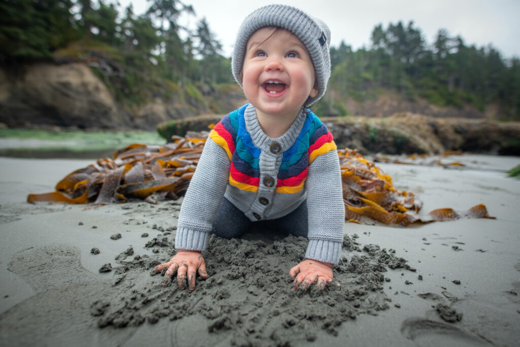 Baby smiling whilst digging in sand on beach with seaweed right behind them and trees & cliffs in background. Photo taken during family roadtrip to Northwestern Edge of Olympic Peninsula. 