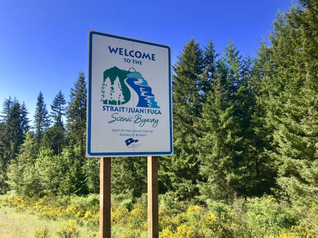 Strait of Juan de Fuca Scenic Byway Welcome Sign in front of trees and blue sky seen during family road trip to Northwestern Edge of Olympic Peninsula.