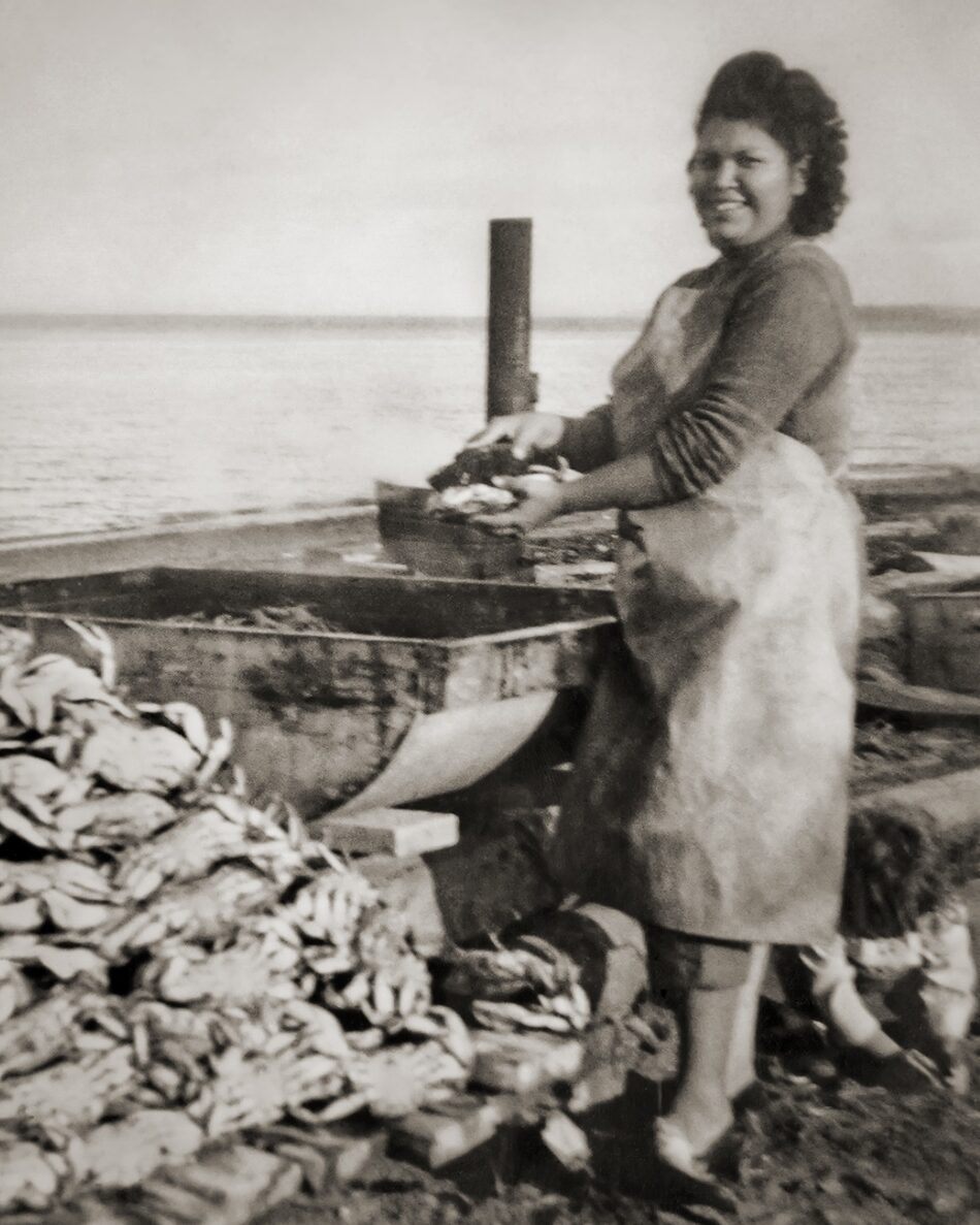 black and white photo of tribe member cleaning crabs with water in the background