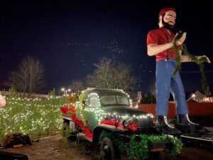 Photo of a entry into the Shelton Holiday Magic Parade. The photo shows an old truck with a giant Paul Bunyan scultpure attached to the front of it. Everything is decorated in white Christmas lights.