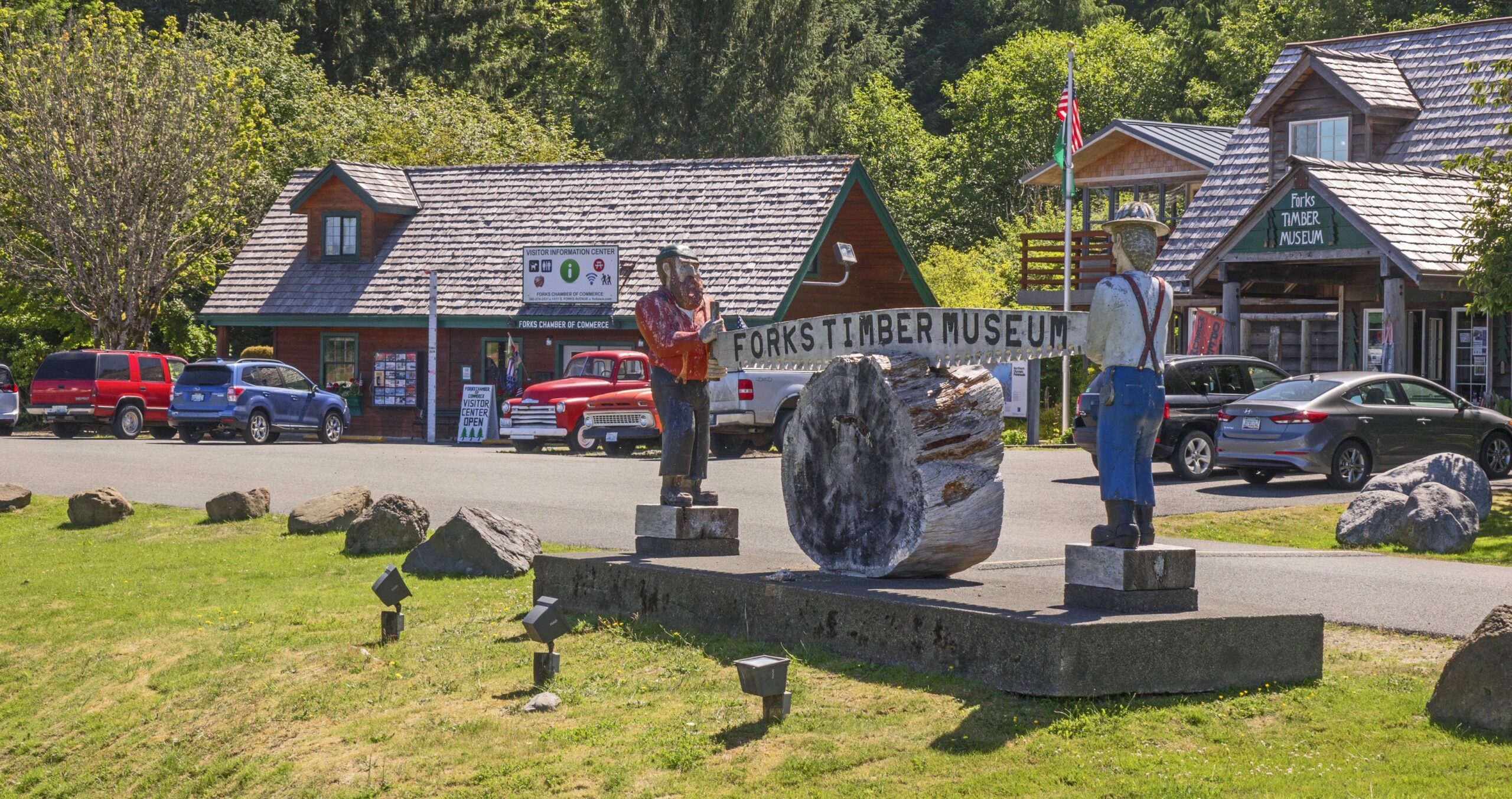 Photo taken outside of Forks Timber Museum in Forks, WA that shows the museum, visitor information center, and wooden statues cutting a log